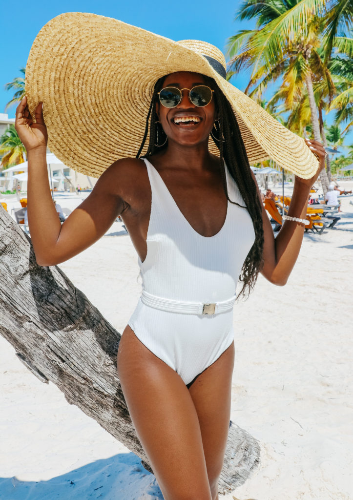 woman smiling and wearing white bathing suit and oversize beach hat